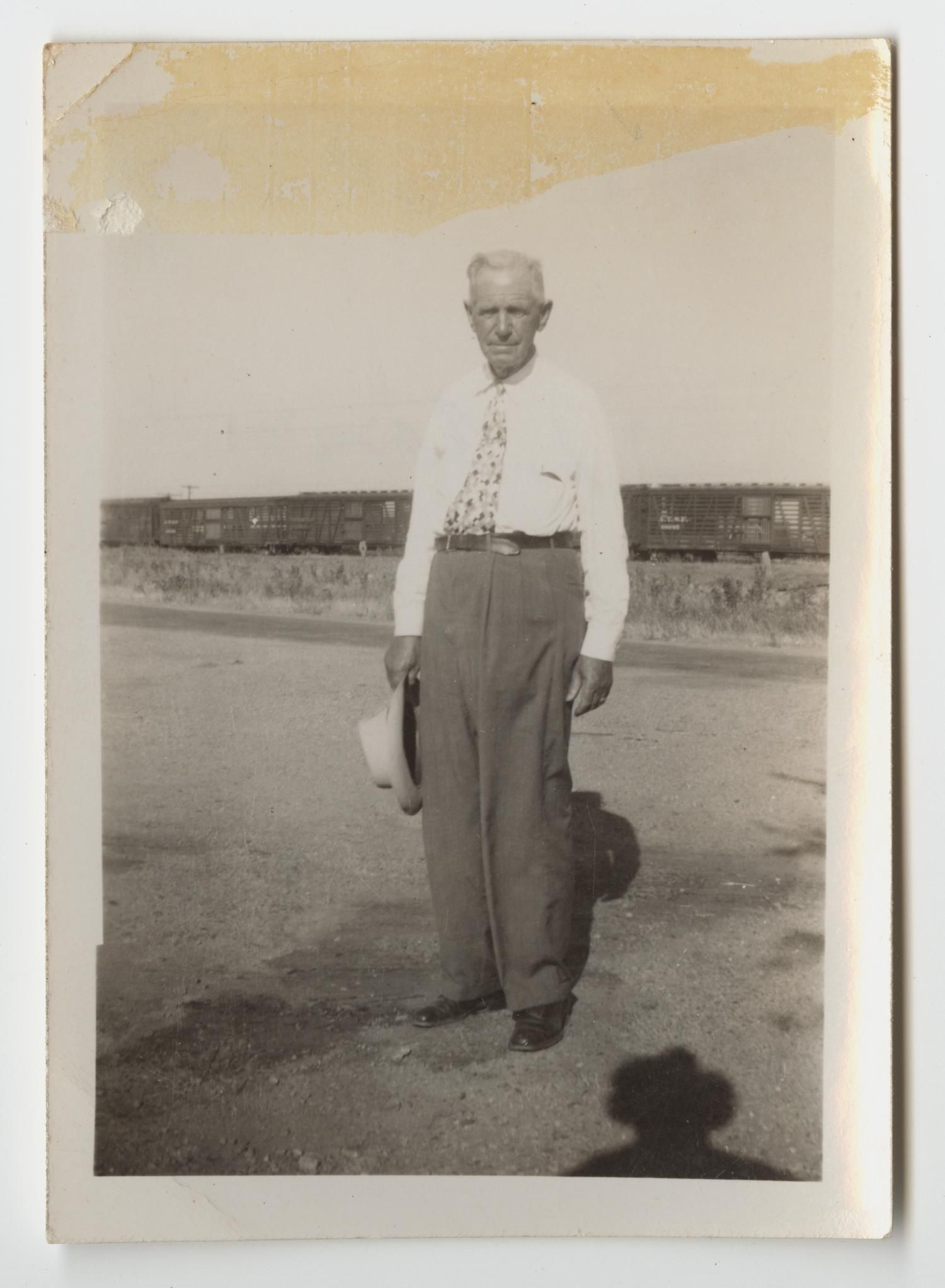 An older man in a white shirt and patterned tie stands on a dirt roadside holding a hat, with a line of railroad boxcars passing behind him.