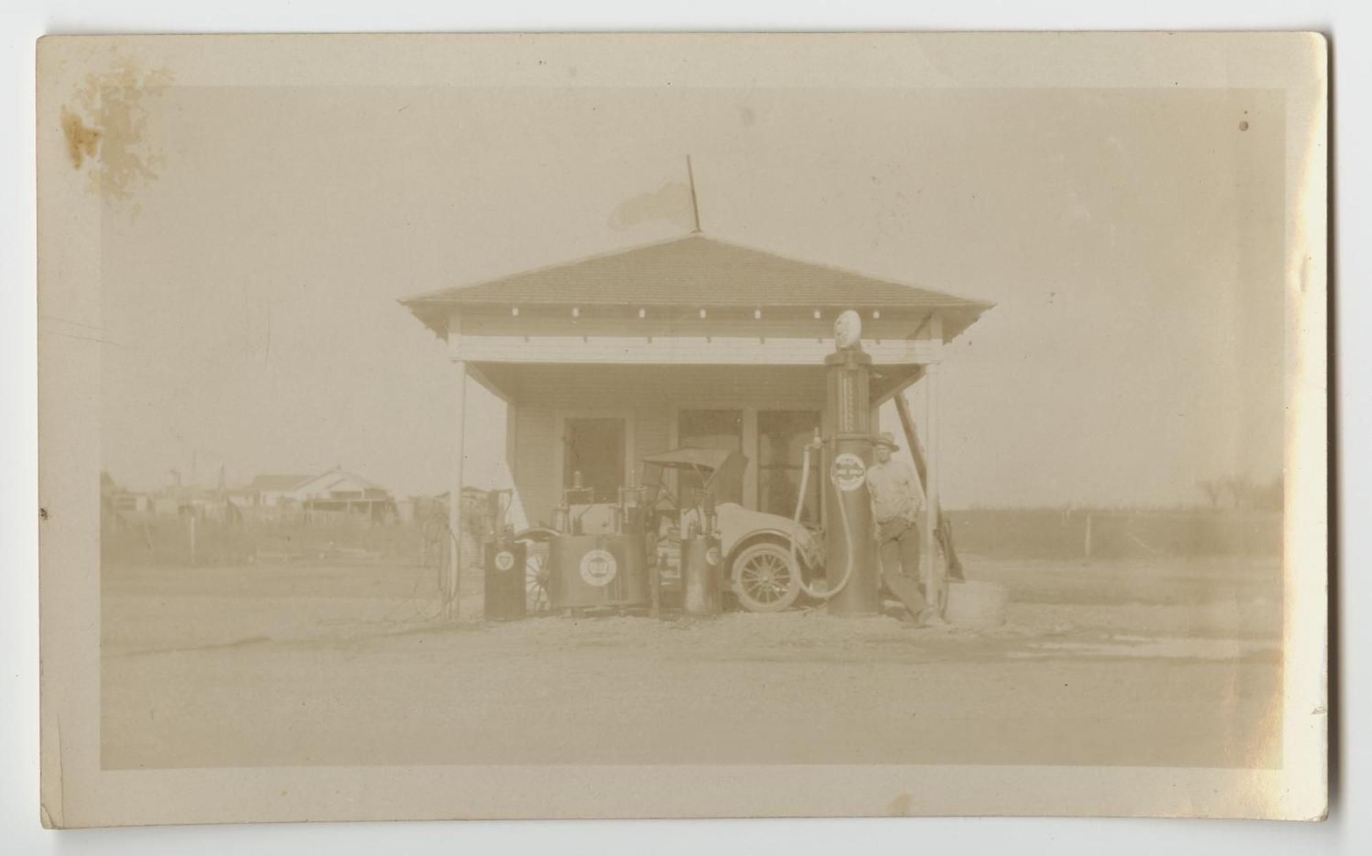 A small filling station with a hipped roof and tall glass-topped gas pumps, a 1920s automobile parked at the pumps, and a man leaning on one pump.