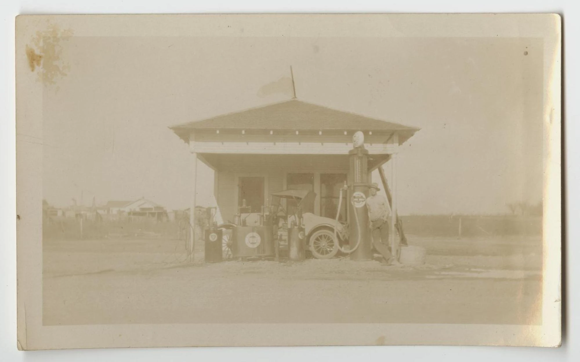 A small filling station with a hipped roof and tall glass-topped gas pumps, a 1920s automobile parked at the pumps, and a man leaning on one pump.