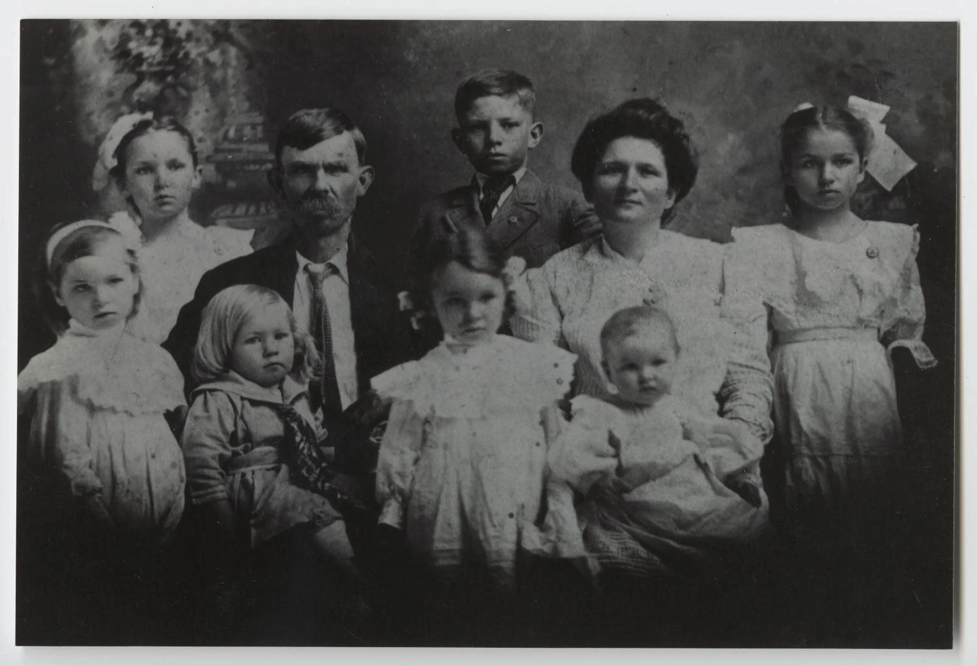 A formal studio portrait of a mustached father and a mother seated among seven children, the girls in white dresses and hair bows.
