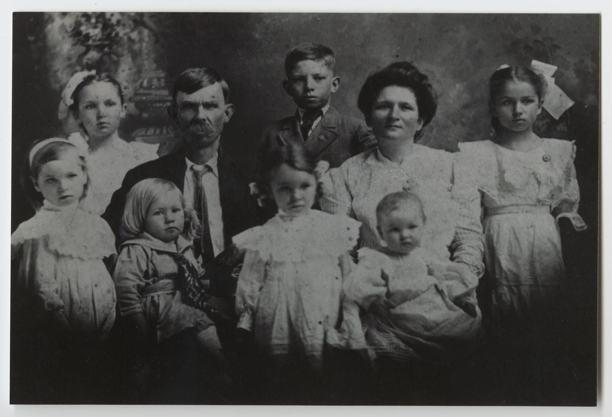 A formal studio portrait of a mustached father and a mother seated among seven children, the girls in white dresses and hair bows.