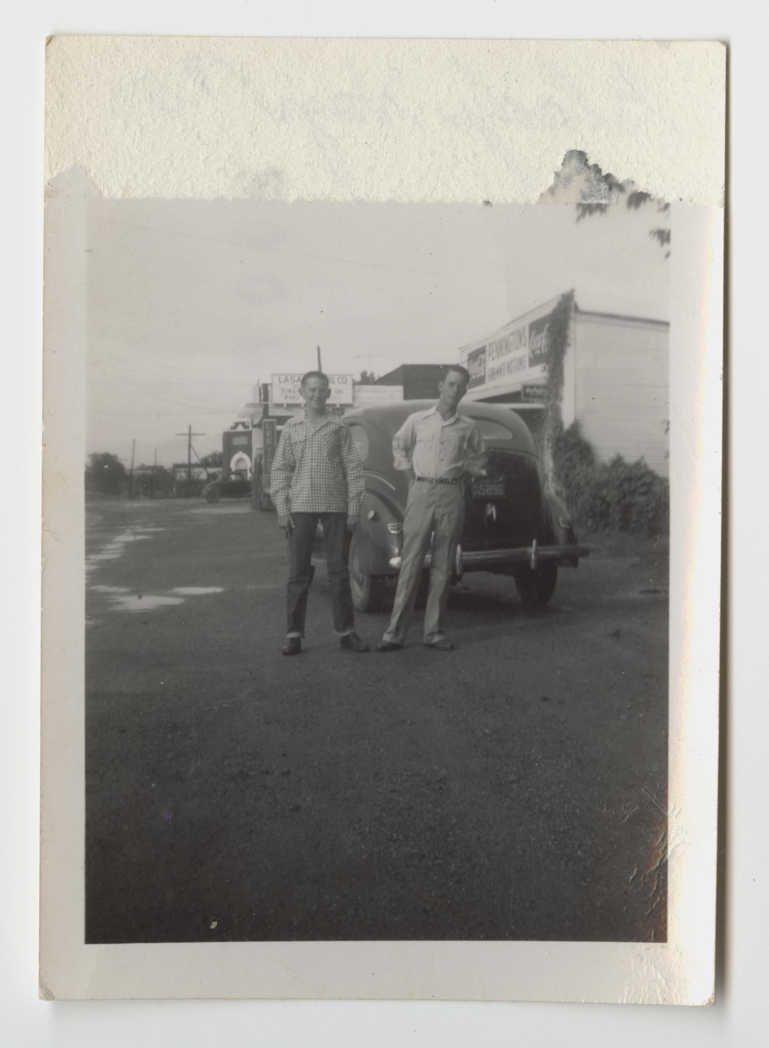 Two young men stand in front of a 1940s car on the gravel shoulder of FM 156, with a row of small storefronts and signs stretching behind them.