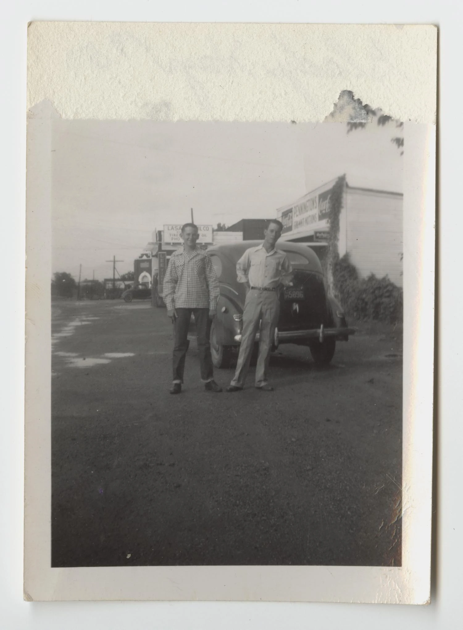 Two young men stand in front of a 1940s car on the gravel shoulder of FM 156, with a row of small storefronts and signs stretching behind them.
