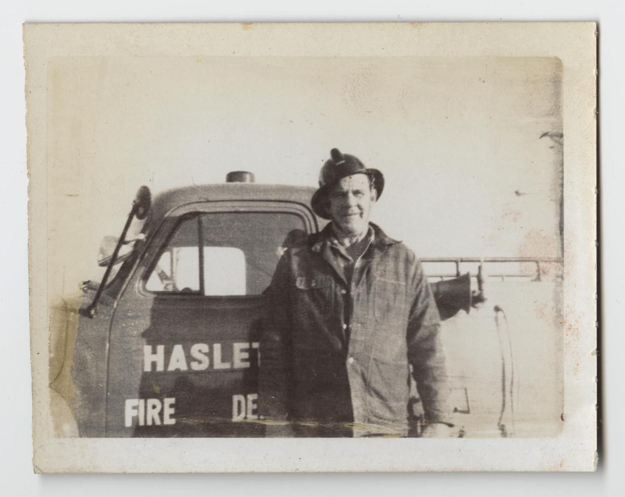 A smiling man in a firefighter's helmet and heavy coat stands beside a fire truck door lettered "Haslet Fire Dept."