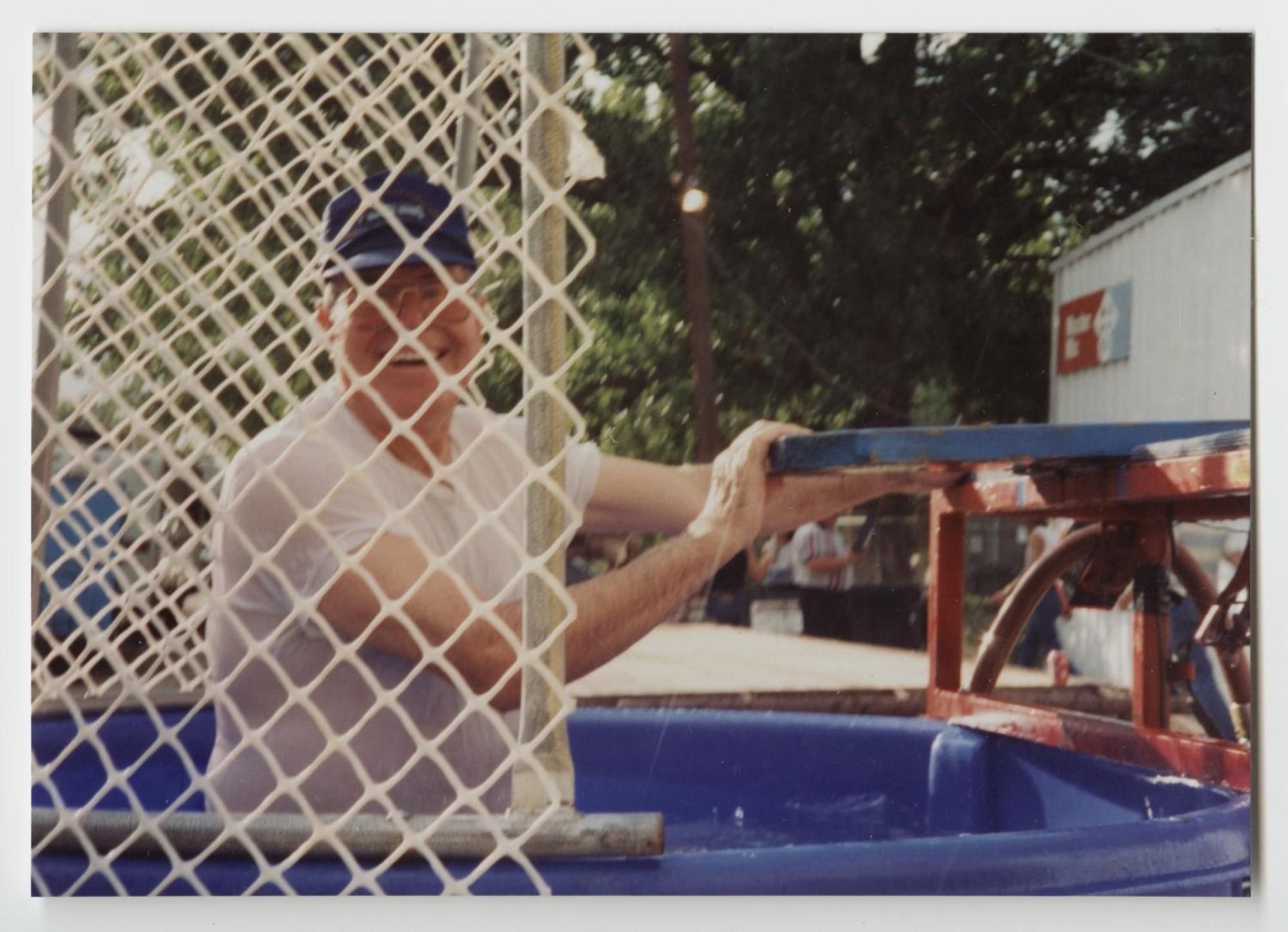 A soaked older man in a cap sits in the water tank of a dunking booth behind a chain-link screen, laughing.