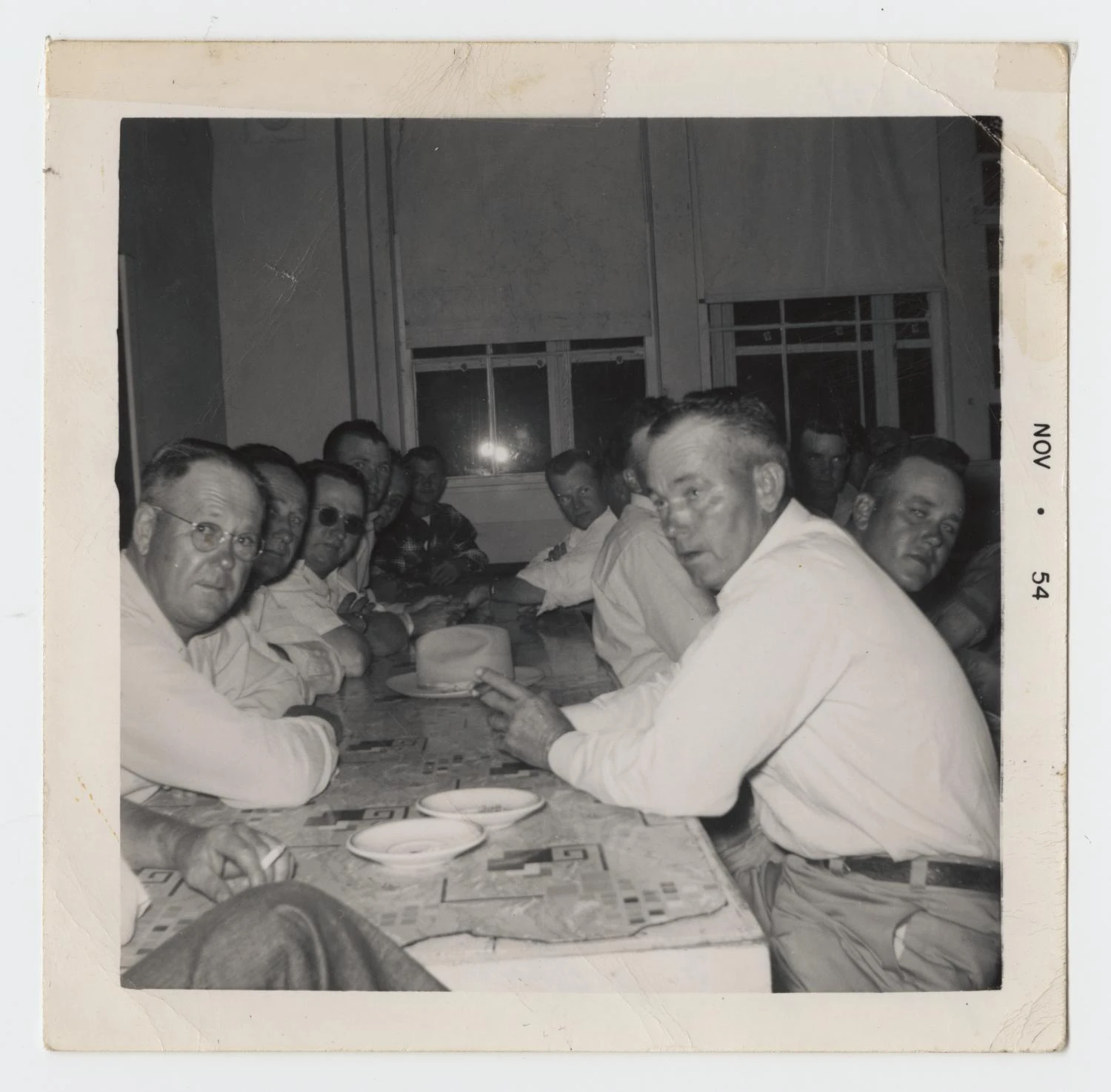 A long table of men in white shirts, several smoking, turn toward the camera during an evening meeting. The print is stamped "Nov 54."