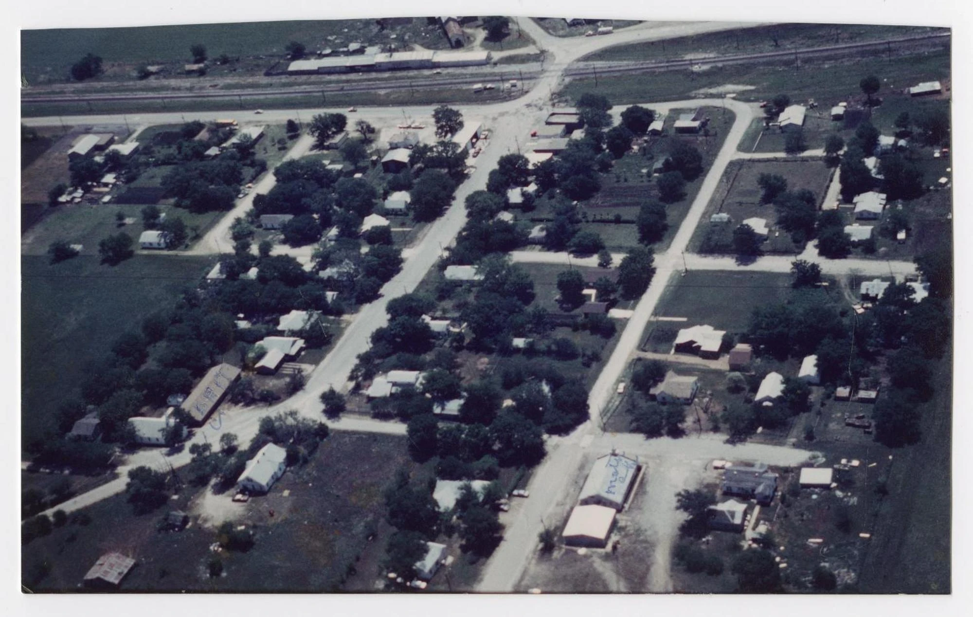 An aerial color photograph of a small town grid: tree-lined streets, scattered houses and low buildings, and a railroad line running across the top.