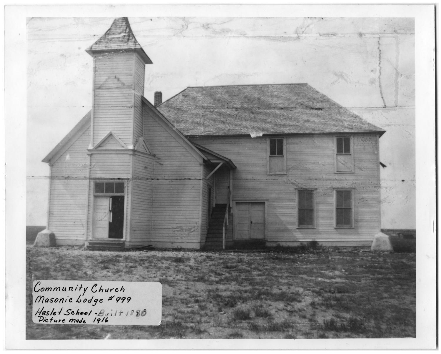 A two-story white clapboard building with a pointed bell tower on the left, an outside staircase, and open prairie around it. A handwritten label reads "Community Church, Masonic Lodge #999, Haslet School, Built 1880, Picture made 1916."