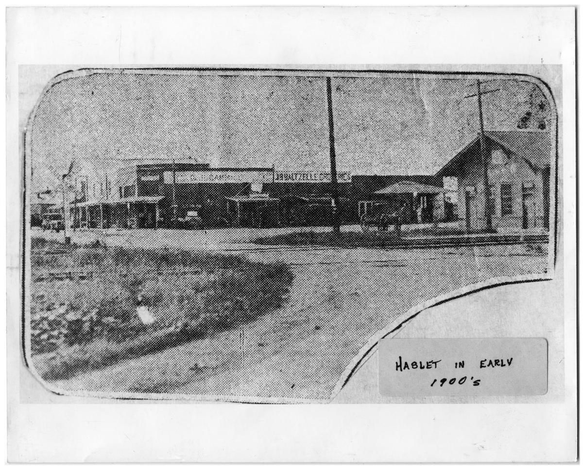 A dirt street curving past a row of false-front stores with signs for Gammill and Baltzelle Groceries, a horse-drawn wagon, utility poles, and a depot-style building at right. A label reads "Haslet in early 1900's."