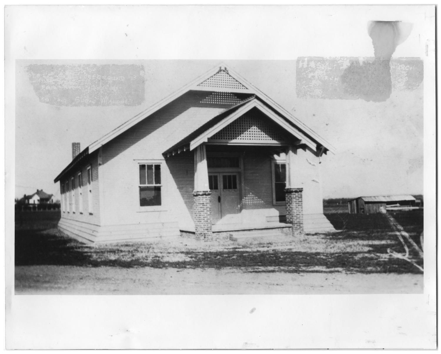 A small one-story white church with a gabled porch on brick columns, a house far in the left background and a barn at right, on flat open ground.