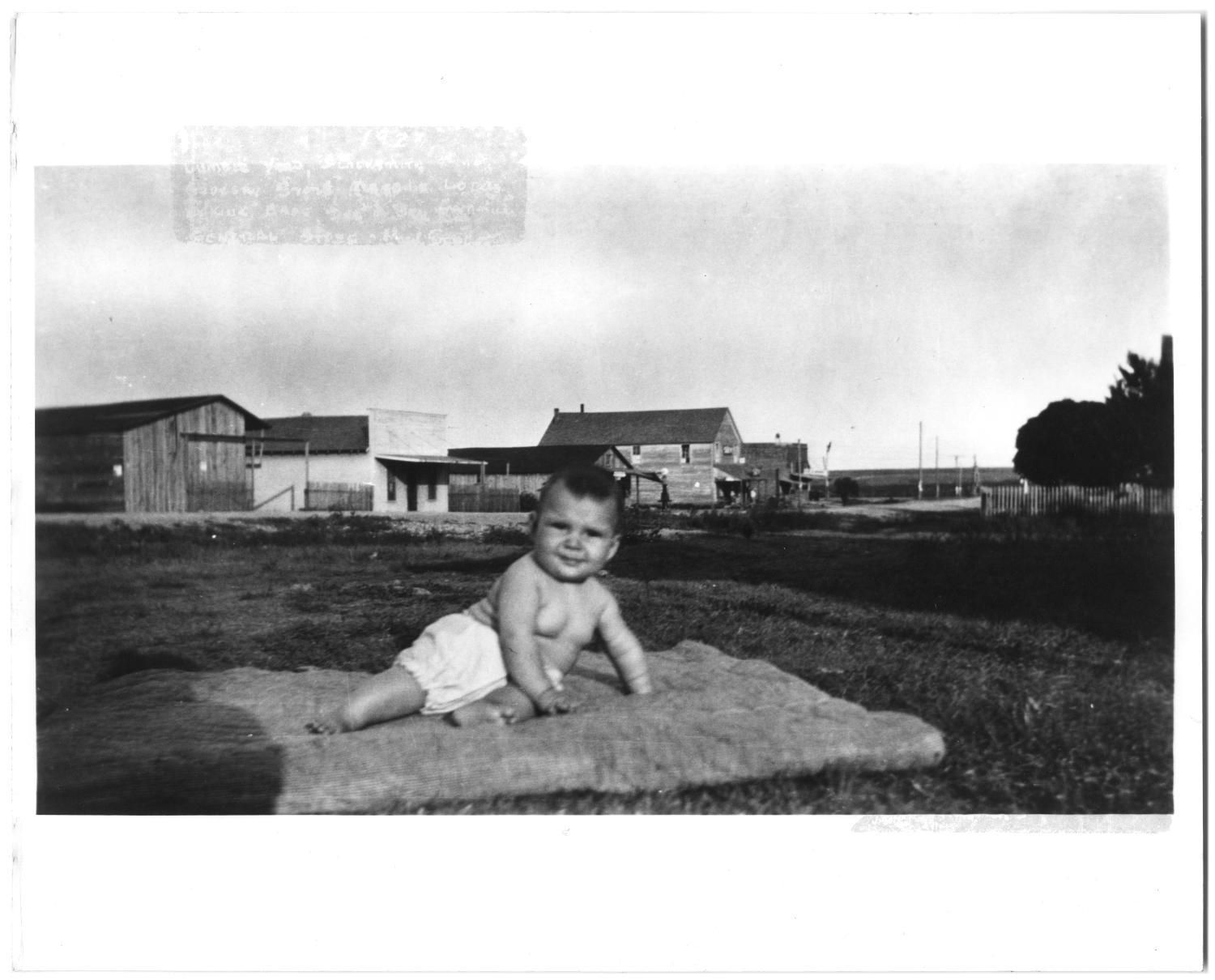 A baby sits on a blanket in a grassy field. Behind her, across the road, is a row of low wooden commercial buildings along Main Street.