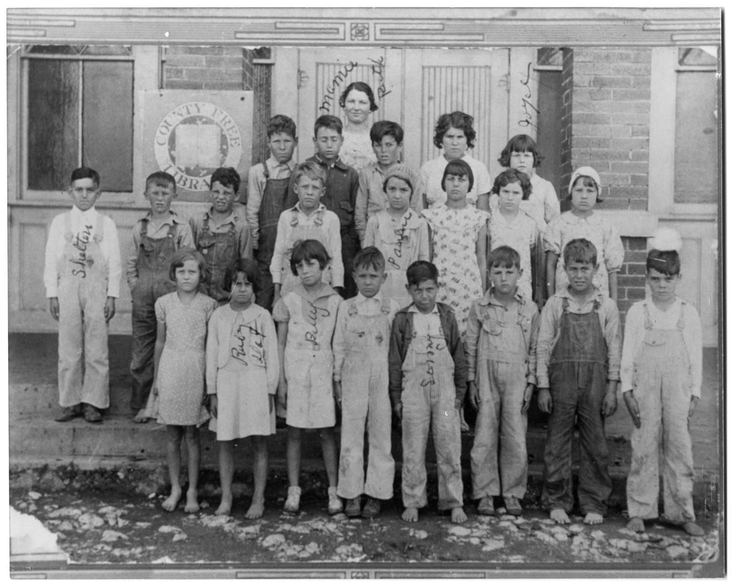 About twenty children in overalls and dresses, several barefoot, stand in rows with their teacher in front of a brick building with a "County Free Library" sign.