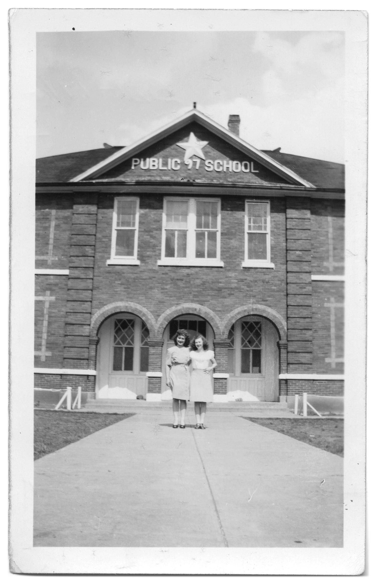 Two young women stand on a long front walk before a two-story brick school with arched doorways and "Public School" and a star lettered in the gable.